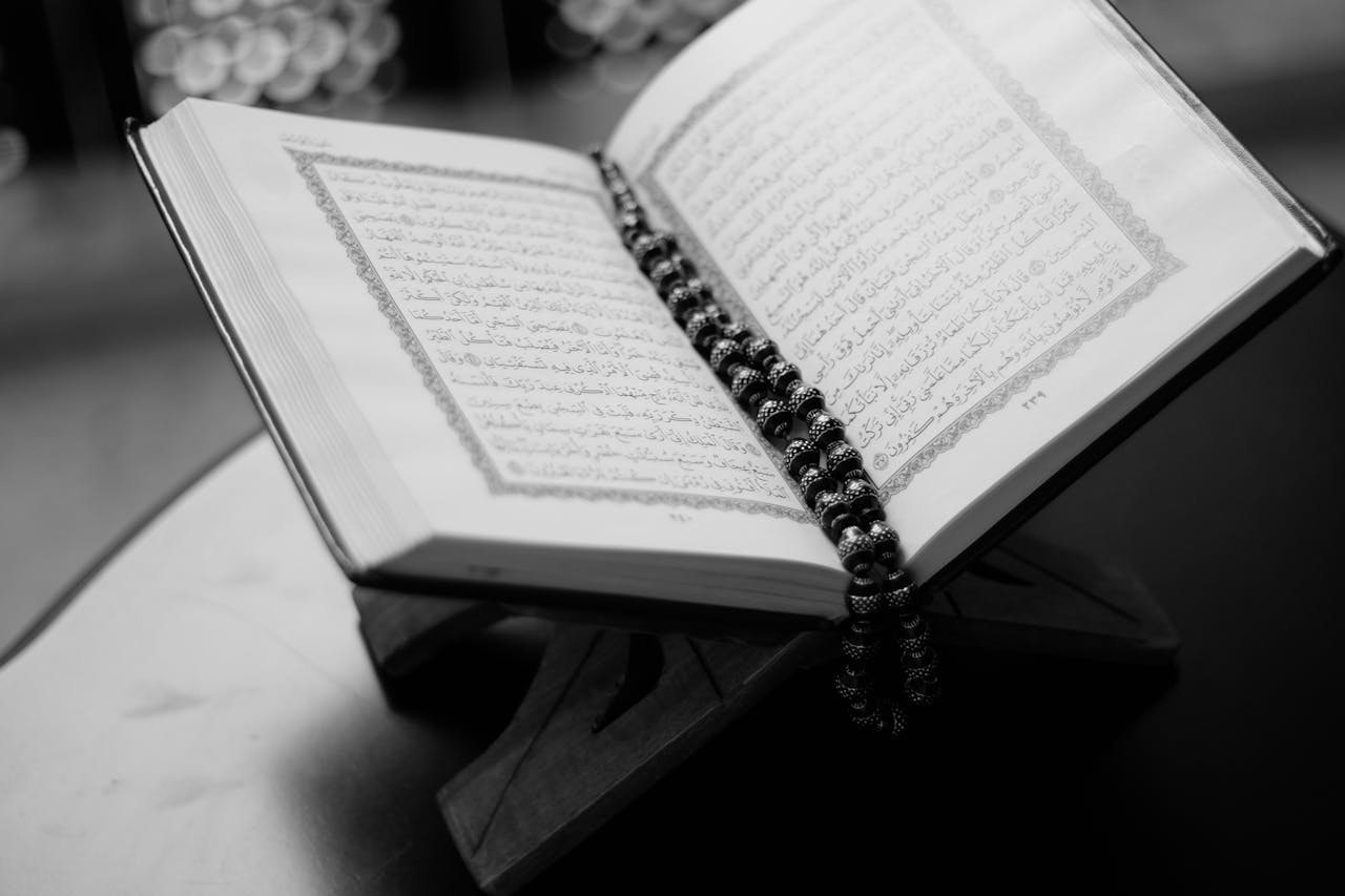 A black and white close-up of an open Quran on a wooden stand with prayer beads, symbolizing Islamic faith and devotion.